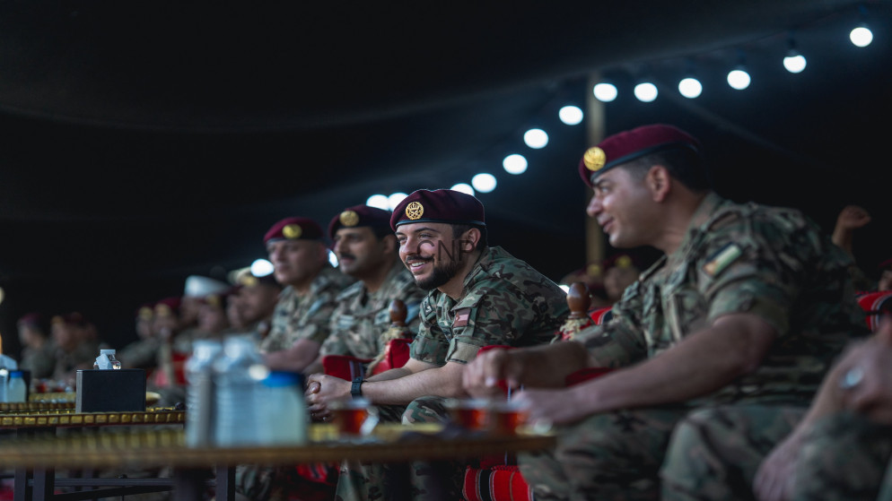 Group of soldiers in camouflage uniforms and maroon berets seated at a long table at a dimly lit event, smiling and watching something off-camera.
