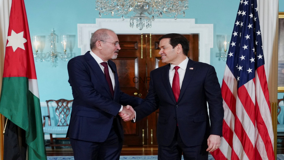 image - Center News Network Two suited men shake hands in a formal room, flanked by the Jordanian and American flags behind them.