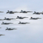 Twelve fighter jets flying in a tight formation across a clear blue sky, with a larger support aircraft at the far right.