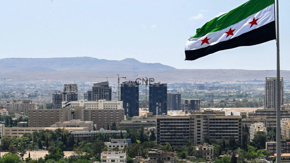 City skyline with a large Syrian flag on a tall pole in the foreground and construction cranes behind, under a clear blue sky and distant hills.