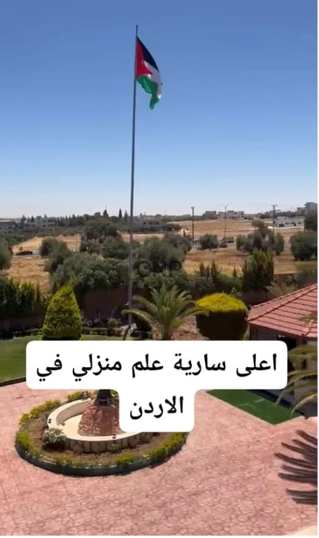 image - Center News Network Tall flagpole with the Jordan flag over a landscaped courtyard; circular planting bed, palm trees, clear blue sky. Arabic caption: أعلى سارية علم منزلي في الأردن.