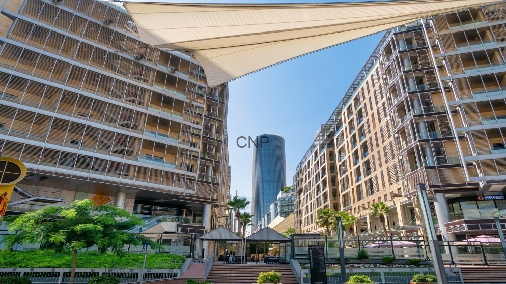 Open-air plaza framed by two curved beige buildings, with a tall blue glass tower in the background and shaded seating areas amid palm trees and greenery.