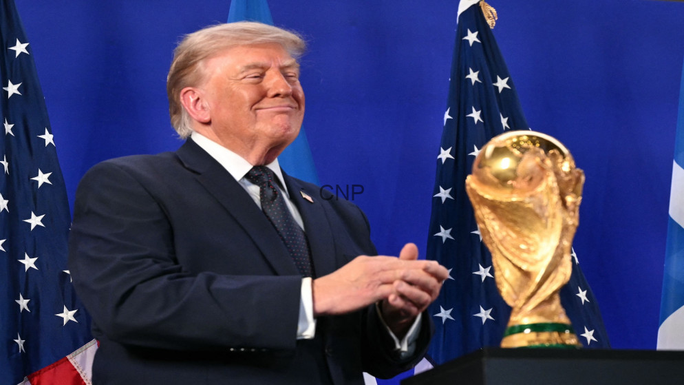 image - Center News Network Donald Trump in a suit, smiling and clapping at a podium, with American flags in the background and a gold trophy nearby.