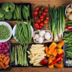 Assorted fresh vegetables arranged on a wooden surface, including broccoli, peas, green beans, cherry tomatoes, peppers, carrots, potatoes, and leafy greens.