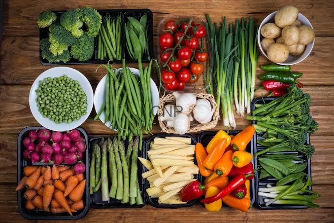 Assorted fresh vegetables arranged on a wooden surface, including broccoli, peas, green beans, cherry tomatoes, peppers, carrots, potatoes, and leafy greens.