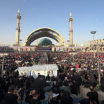 Large crowd gathered at a mosque plaza with a green-domed shrine and tall minarets during a religious event.