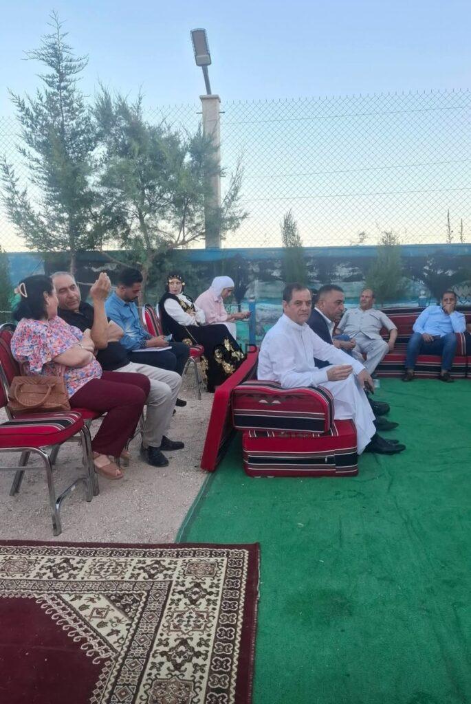 Group of people seated in a semi-circle on red cushioned chairs at an outdoor gathering, with green carpet and decorative rugs, chatting in daylight.