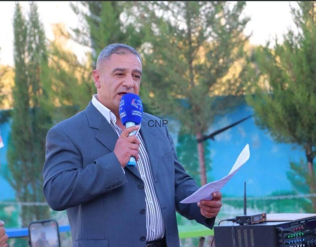 Man in a gray suit speaks into a blue handheld microphone outdoors, holding a sheet of paper on a sunny day.