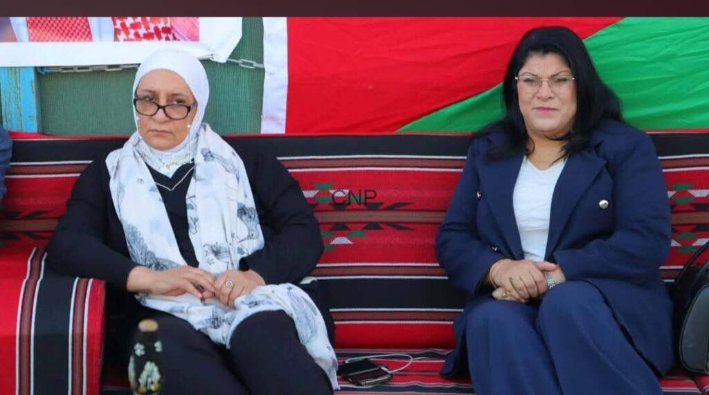 Two women sit side by side at a cultural event on a red-and-black woven bench, one in a white headscarf and glasses and the other in a navy suit, both looking toward the camera.