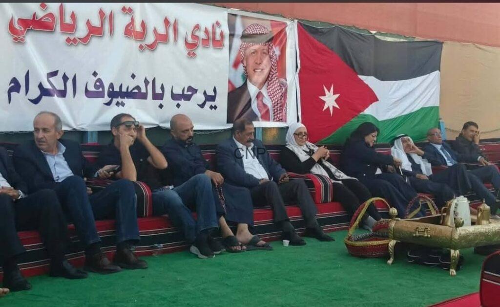 mas4 - Center News Network Group of men and women seated on a long red sofa at a Jordanian event, with an Arabic banner and the Jordan flag in the background.