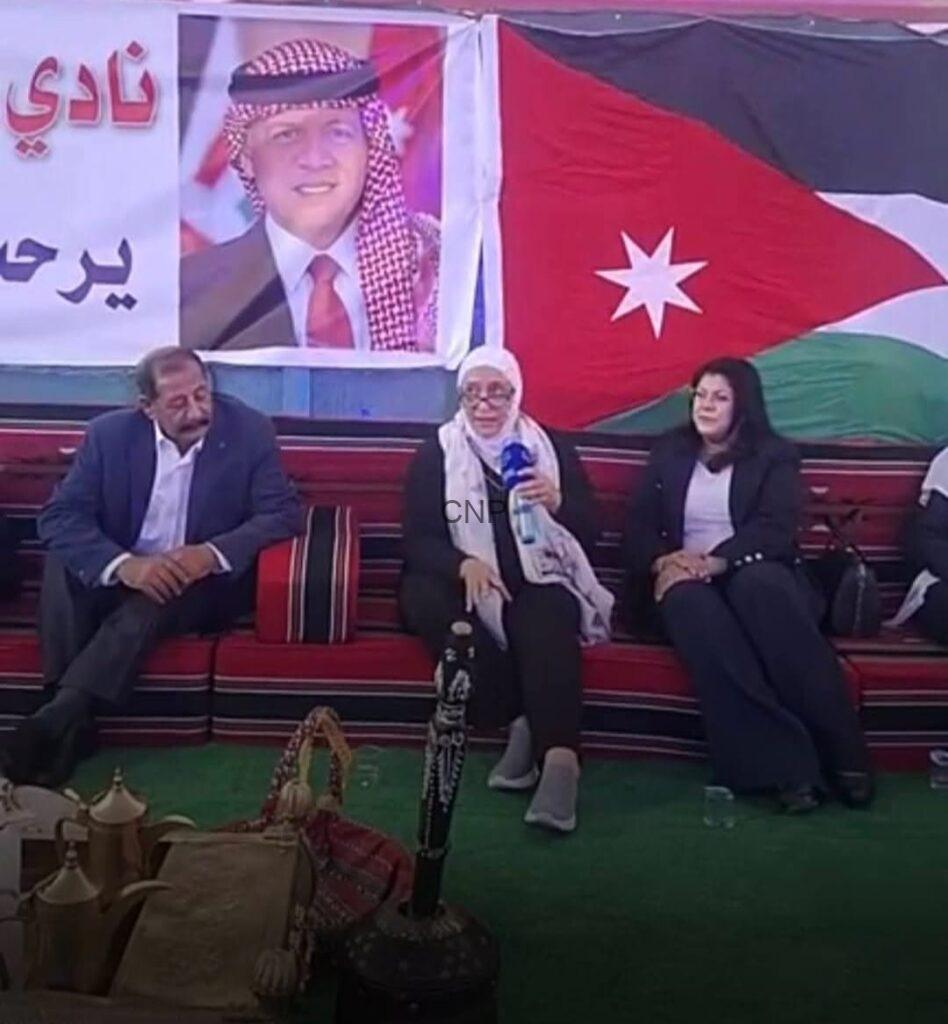 Three people seated on a red bench at a cultural event, with Jordanian flag and a portrait banner behind them.