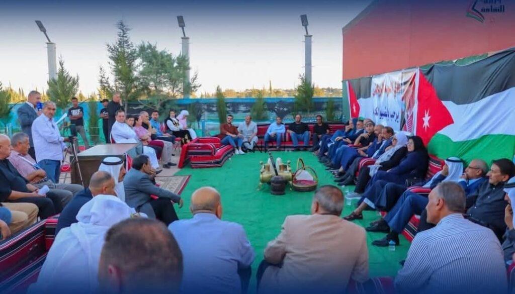 Group of people seated in a circle on a green carpet outdoors for a community gathering, baskets in the center and a Jordan flag banner in the background.