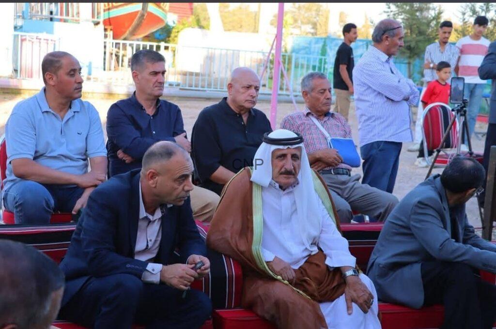 Group of men seated outdoors at an event, one in traditional Middle Eastern dress with a white headscarf and brown cloak.