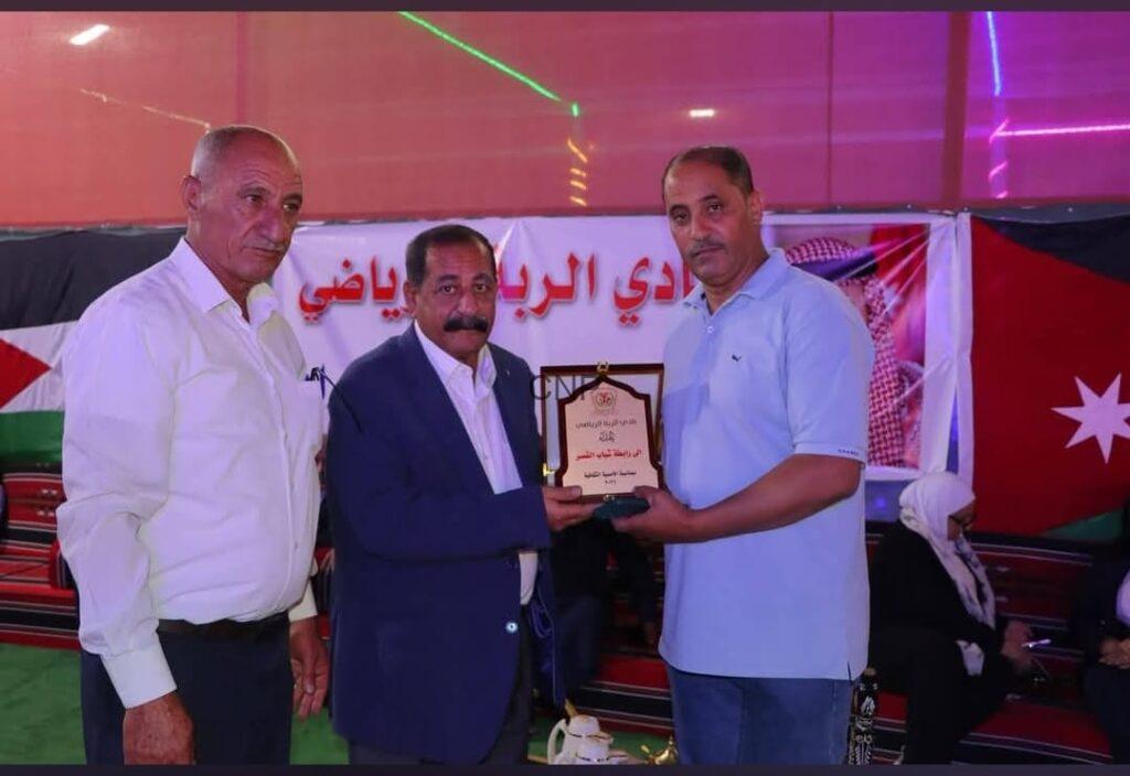 mas8 - Center News Network Three men on a stage during an award ceremony; one man in a suit presents a plaque to another in a light blue polo, with an Arabic banner behind them.