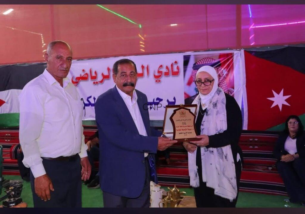 Three people on a stage during an award ceremony; a man in a suit hands a plaque to a woman wearing a hijab and glasses, with banners and flags in the background.