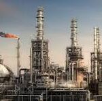 Oil refinery complex with tall smokestacks, distillation towers, and a network of pipes against a clear sky