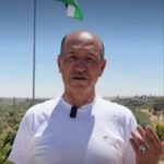 Man wearing a white t-shirt with a lapel microphone speaking outdoors, Jordan flag flying on a pole behind him.