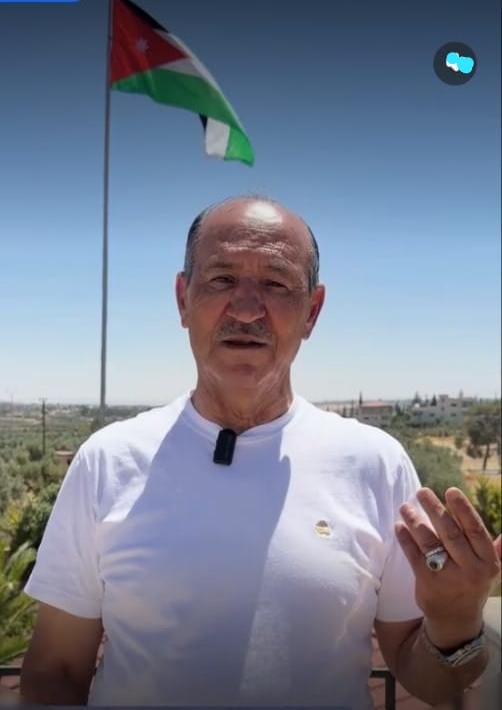 Man wearing a white t-shirt with a lapel microphone speaking outdoors, Jordan flag flying on a pole behind him.