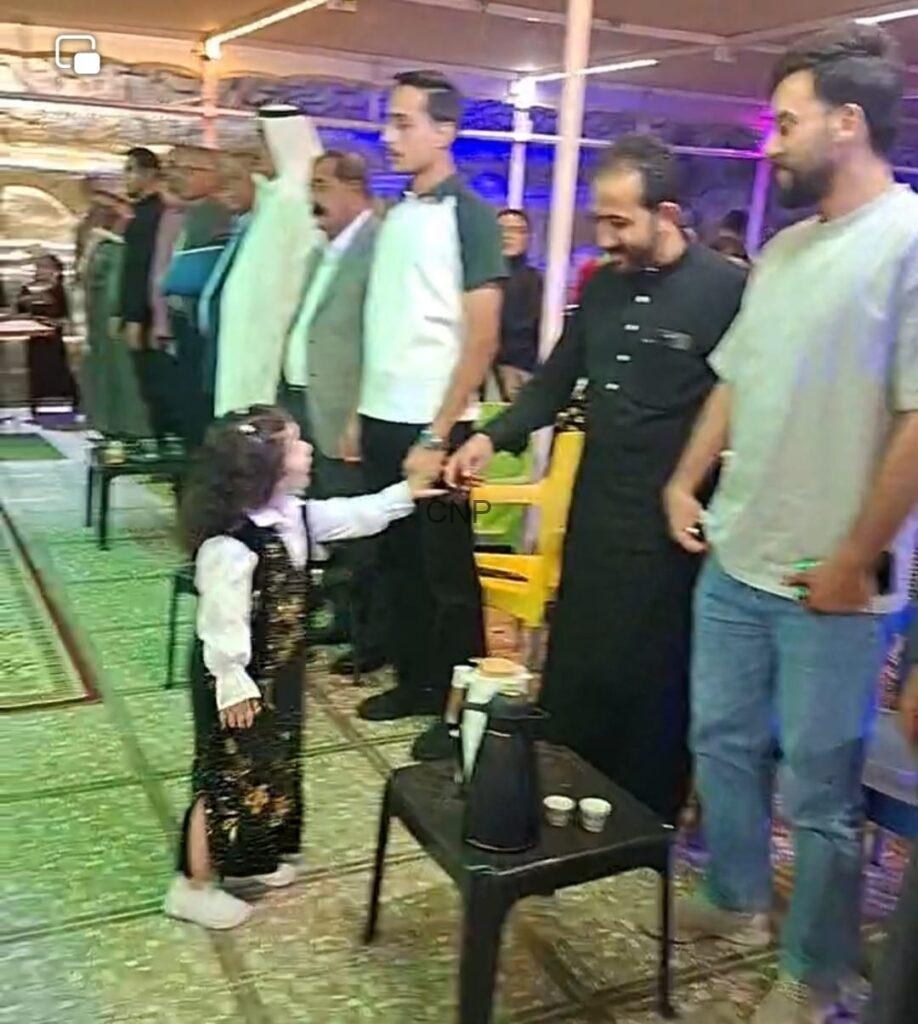Young girl in a sparkly vest shakes hands with a man in black as others line up during a social gathering in a decorated hall.