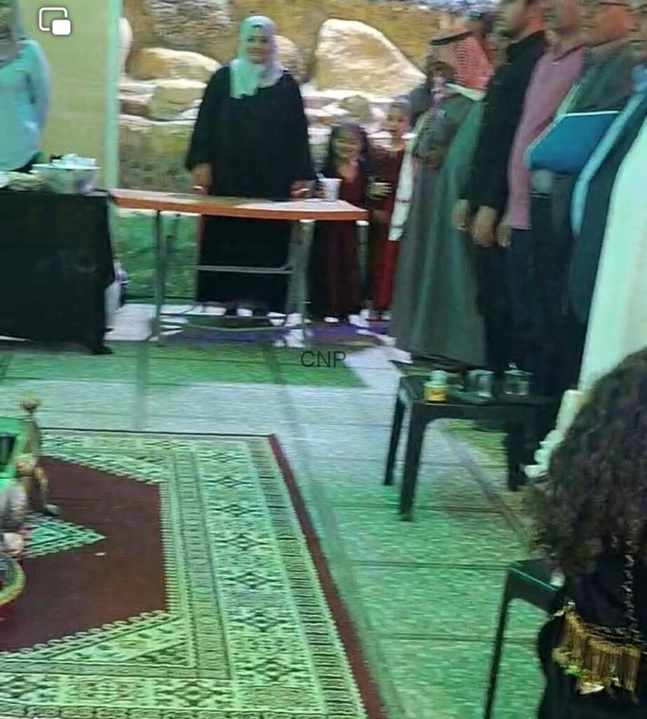 to3 - Center News Network Group of people standing in a room around a wooden table with cups, on a green patterned rug, likely a cultural gathering or ceremony.