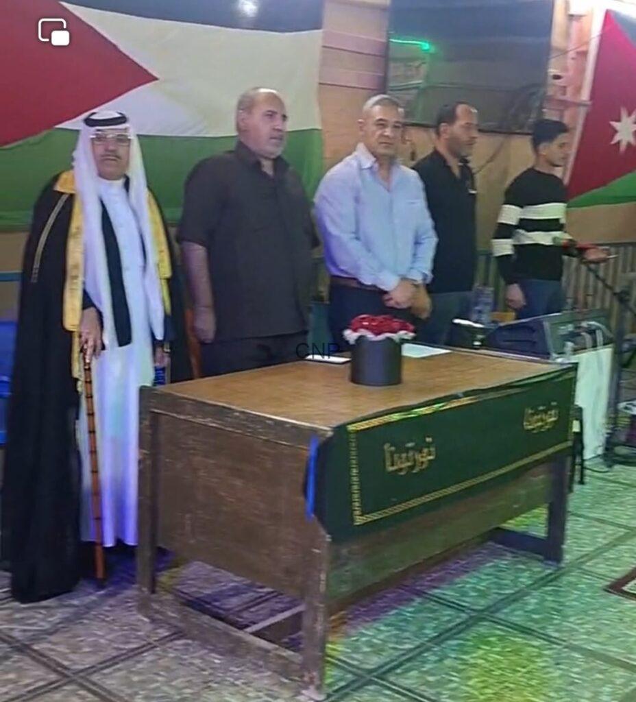 A funeral scene with five men standing beside a wooden coffin draped in a green cloth with gold trim and Arabic inscription, against a backdrop with flags.