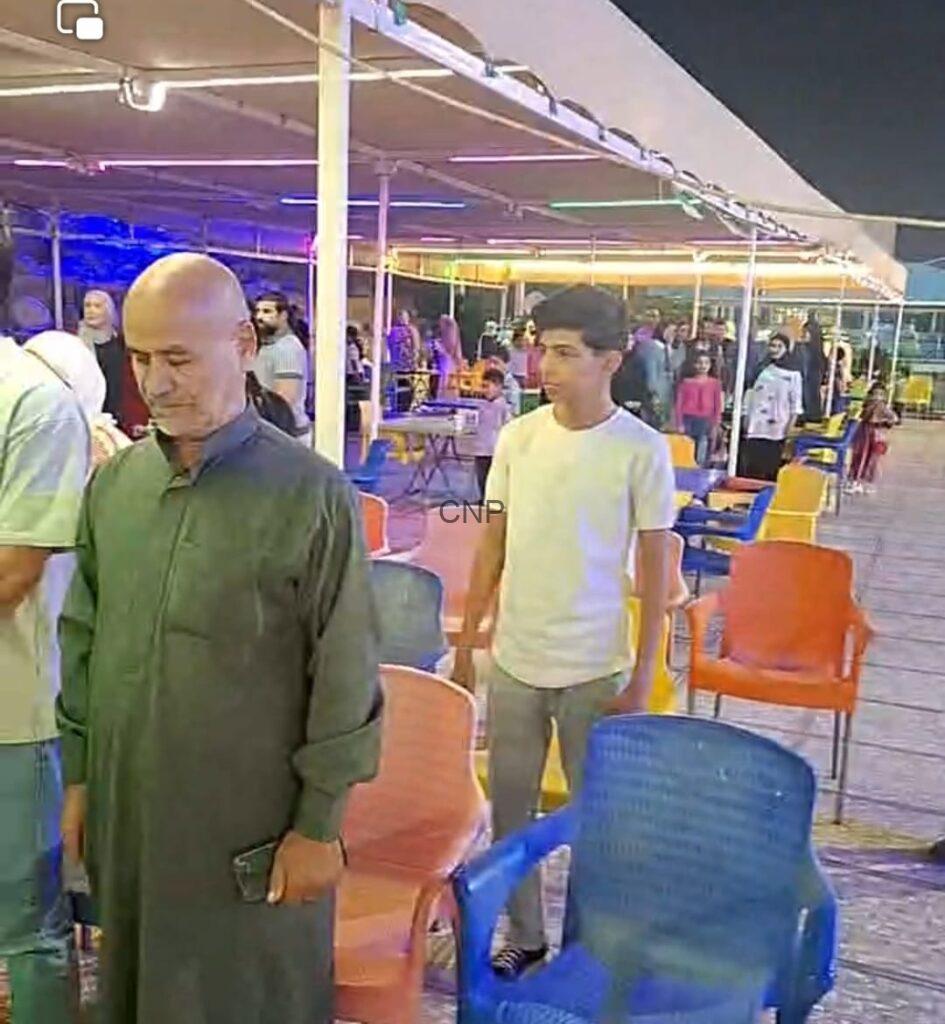 to5 - Center News Network Bald man in a green traditional kurta stands beside colorful plastic chairs at a nighttime outdoor event, holding a phone.