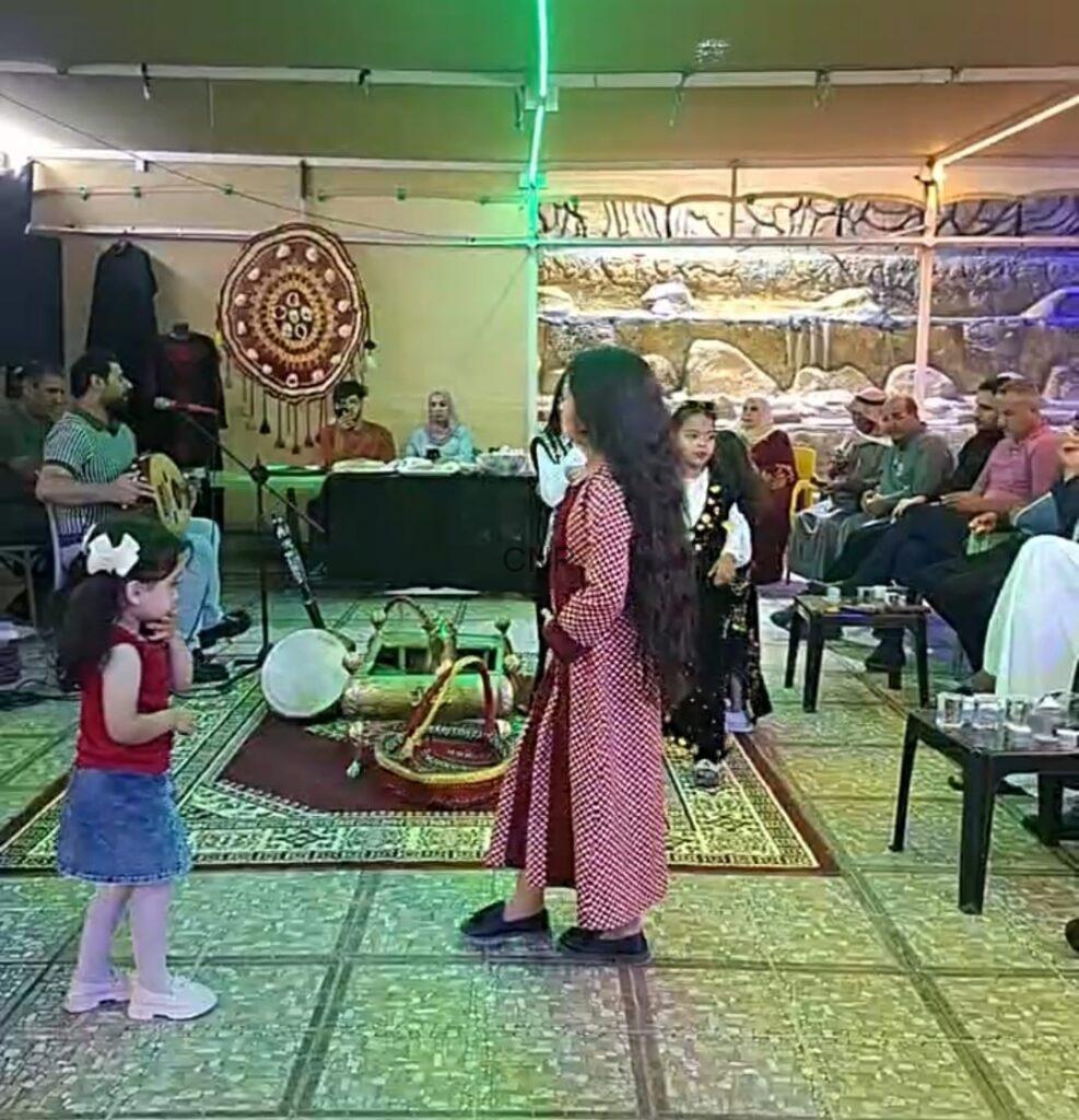 Girl in a red patterned dress dances on a decorative rug while a live band and seated guests watch in a cultural gathering tent.