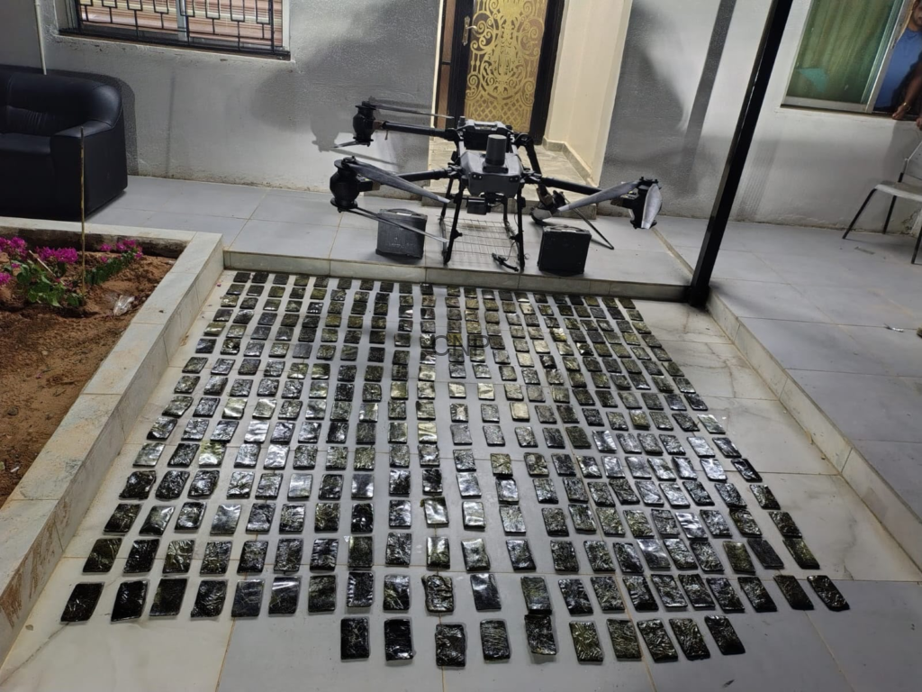 image - Center News Network Drone rests on tiled patio beside a doorway, with hundreds of small rectangular black packages laid out in neat rows on the floor for inspection or inventory.