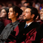 Audience seated at a formal ceremony; a woman in a gray patterned blouse and two men in suits and academic robes are attentively watching the stage.