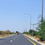 Long two-lane road stretching into the distance, with yellow edge lines and tall streetlights on both sides, a vehicle ahead, clear blue sky and dry brush along the shoulders.
