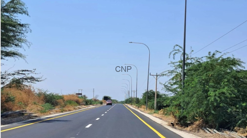 image - Center News Network Long two-lane road stretching into the distance, with yellow edge lines and tall streetlights on both sides, a vehicle ahead, clear blue sky and dry brush along the shoulders.