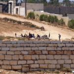 Group of people sitting around a table on a dusty slope behind a low stone wall; a man stands to the right.