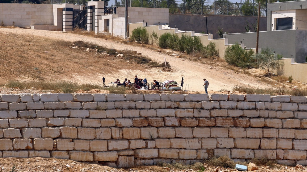 image - Center News Network Group of people sitting around a table on a dusty slope behind a low stone wall; a man stands to the right.
