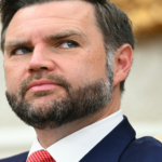 Close-up portrait of a man with a beard in a navy suit and red tie, looking to the left.