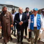 Group of men walking together through a desert camp with tents, led by a man in a dark suit and others in traditional attire and a blue-vested volunteer.