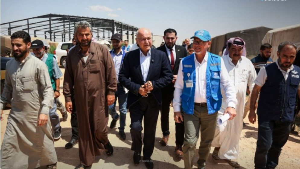 image - Center News Network Group of men walking together through a desert camp with tents, led by a man in a dark suit and others in traditional attire and a blue-vested volunteer.
