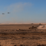 Armored tanks advance across a dusty desert battlefield with helicopters flying overhead.