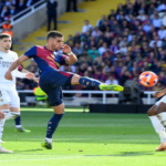 Barcelona player in blue and maroon kicking the ball while Real Madrid players in white close in during a crowded match on a sunny day.