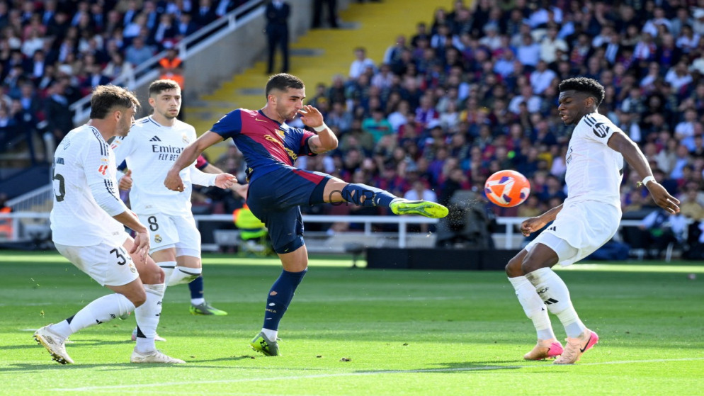 Barcelona player in blue and maroon kicking the ball while Real Madrid players in white close in during a crowded match on a sunny day.