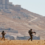 Two soldiers in brown uniforms and helmets patrol a dry hillside with rifles as a town climbs the hill in the background.