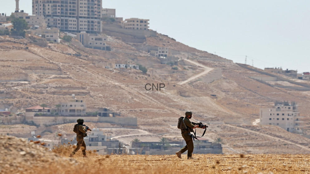 Two soldiers in brown uniforms and helmets patrol a dry hillside with rifles as a town climbs the hill in the background.