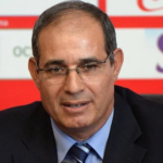 Close-up of a middle-aged man in a dark suit and blue striped tie, wearing glasses, speaking at a press conference with a red backdrop and logos behind him.