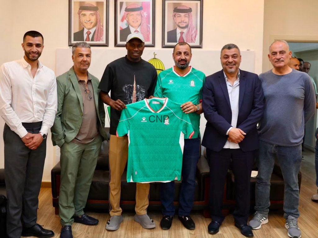 image - Center News Network Seven men standing indoors, smiling, while one holds a green sports jersey in front of them.