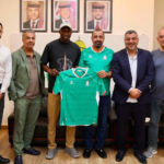 Seven men standing indoors, smiling, while one holds a green sports jersey in front of them.