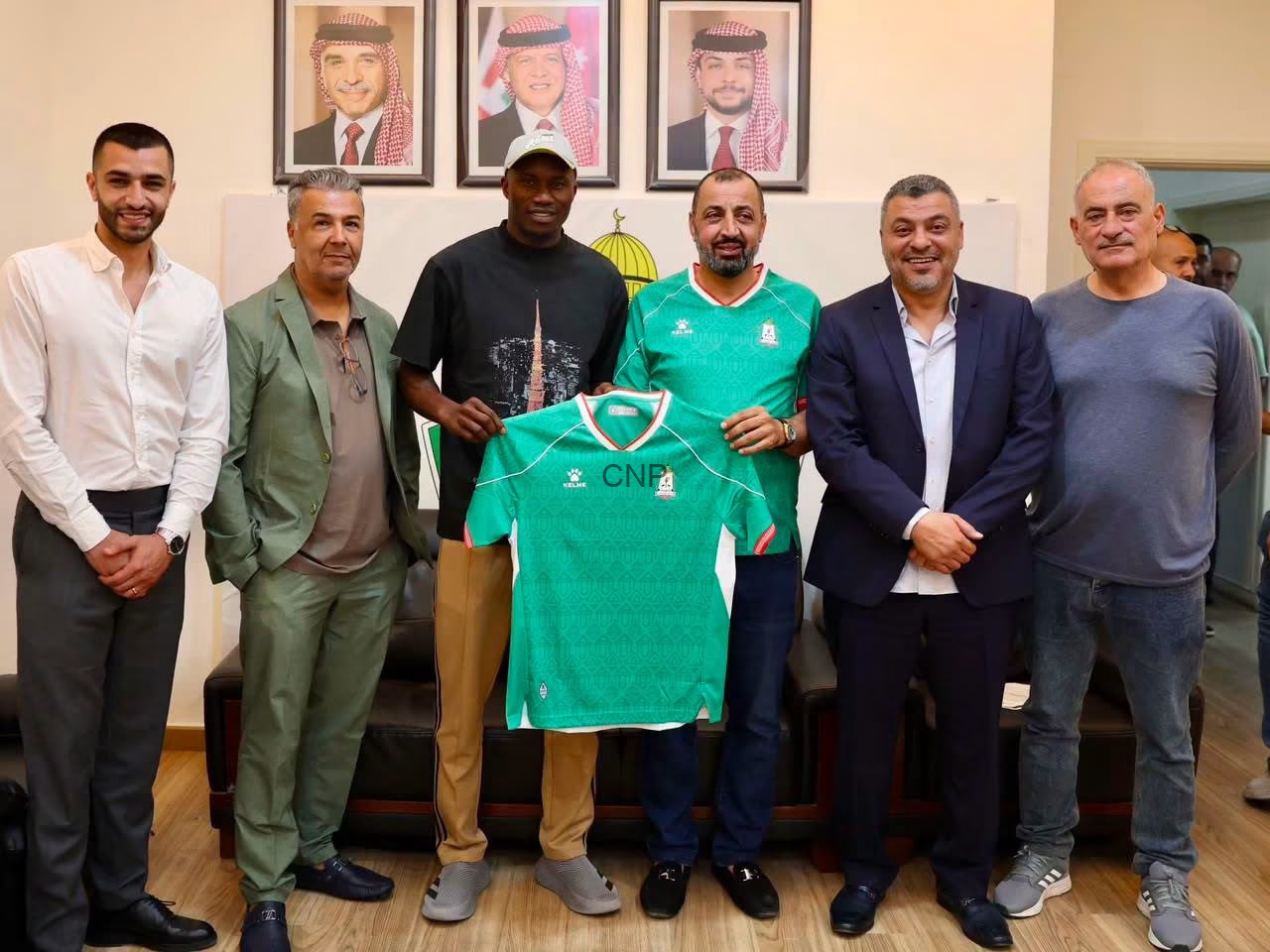image - Center News Network Seven men standing indoors, smiling, while one holds a green sports jersey in front of them.