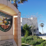 Stone arch and pillar show the Al-Balqa Applied University crest on campus, with trees and a clear blue sky.
