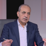 Middle-aged man in a navy pinstripe suit and white shirt sits on a TV talk-show set, gesturing with his hands as he speaks (watermark 'CNP' on chest).