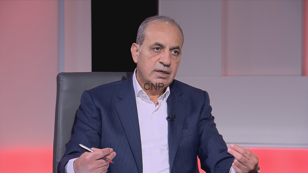 Middle-aged man in a navy pinstripe suit and white shirt sits on a TV talk-show set, gesturing with his hands as he speaks (watermark 'CNP' on chest).