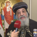 Elderly religious leader in ornate black robes and a rounded headdress being interviewed, holding a red microphone with a religious painting behind him.
