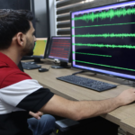 Man in a red shirt sits at a desk, watching a large monitor displaying green audio waveforms.
